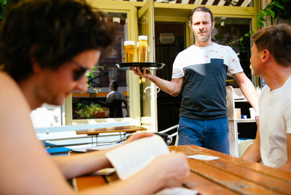 Ein Kellner serviert einem Gast Bier auf einer Café-Terrasse, während eine Person liest.