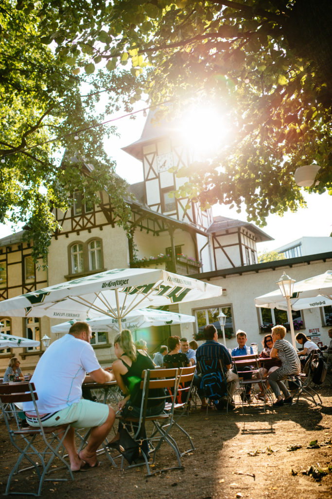 Menschen sitzen entspannt unter grünen Bäumen bei Sonnenschein in einem Außencafé.