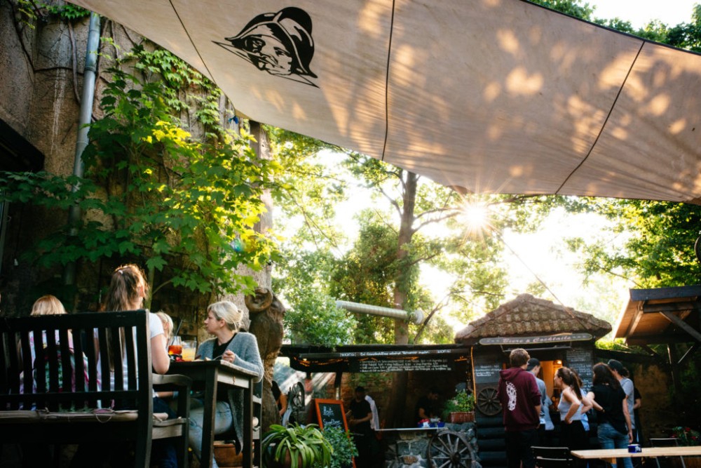Menschen sitzen im Freien in einem Restaurant unter einem Sonnenschirm aus Stoff, Sonnenlicht strahlt zwischen den Blättern durch.