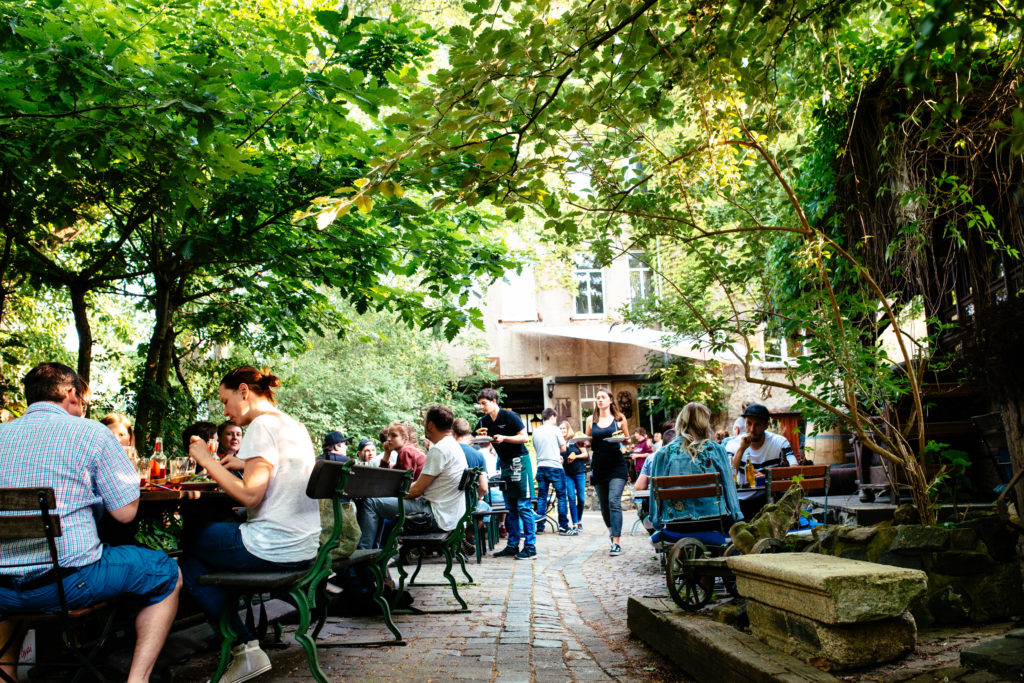 Personen sitzen an Tischen im Freien eines Cafés, umgeben von üppigem Grün und Steindekorationen.