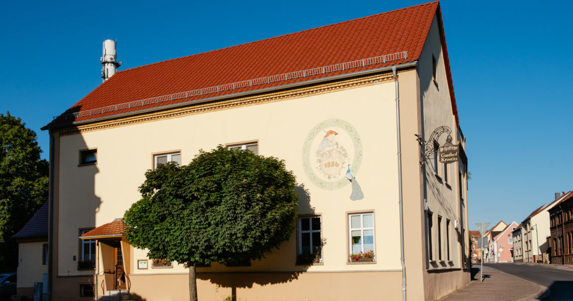 Ein zweistöckiges Gebäude mit rotem Ziegeldach und Werbung an der Fassade steht an einer Straßenecke.