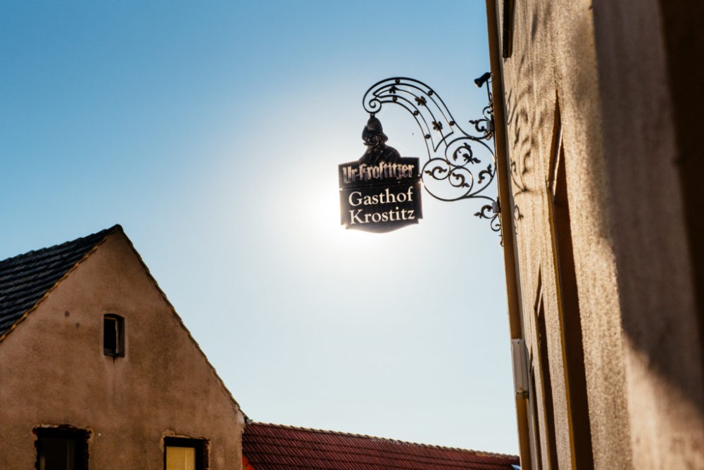 Ein Eisenschild mit Verzierungen hängt an einer Hauswand vor blauem Himmel.