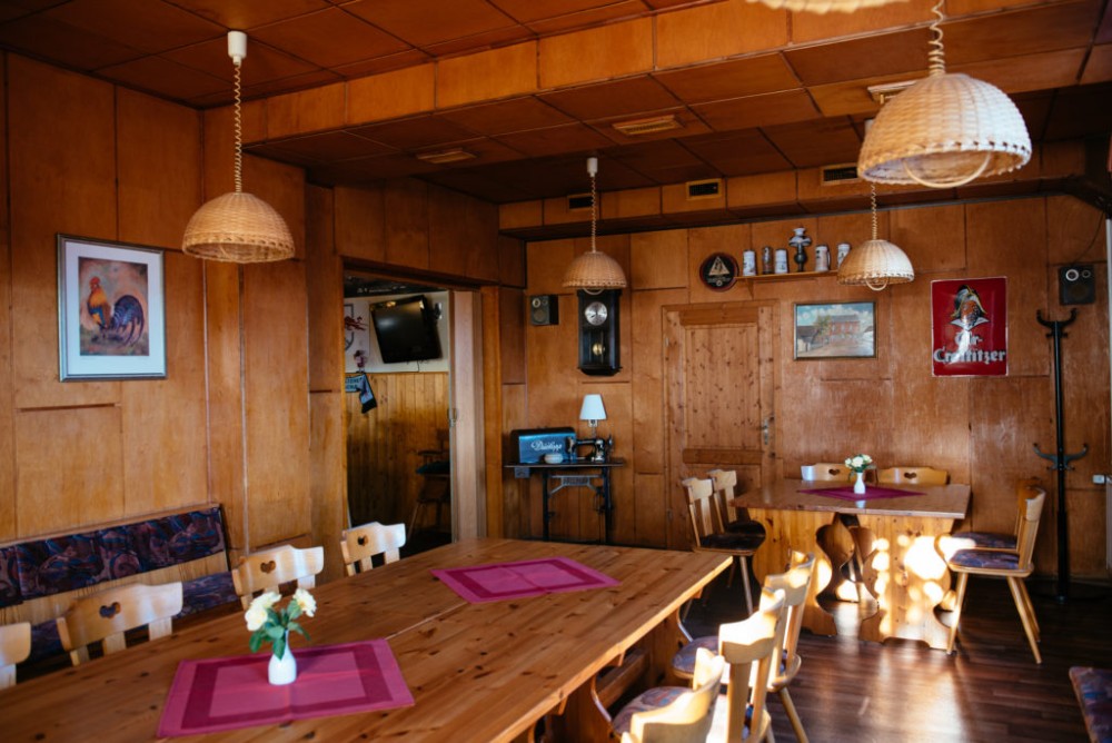 Eine hölzerne Gaststube mit rustikalen Tischen und Körbchenlampen. Wände und Tische zeigen ländliche Dekoration und Kunst.