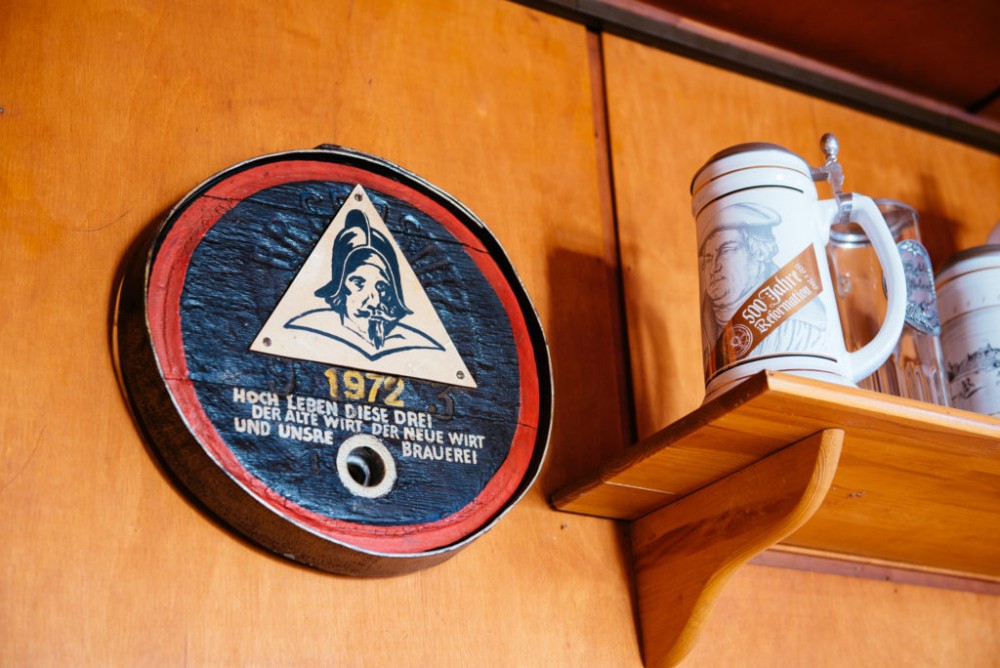 Ein rundes Bierfassschild mit Dreieckslogo hängt neben Bierkrügen an einer Wand.