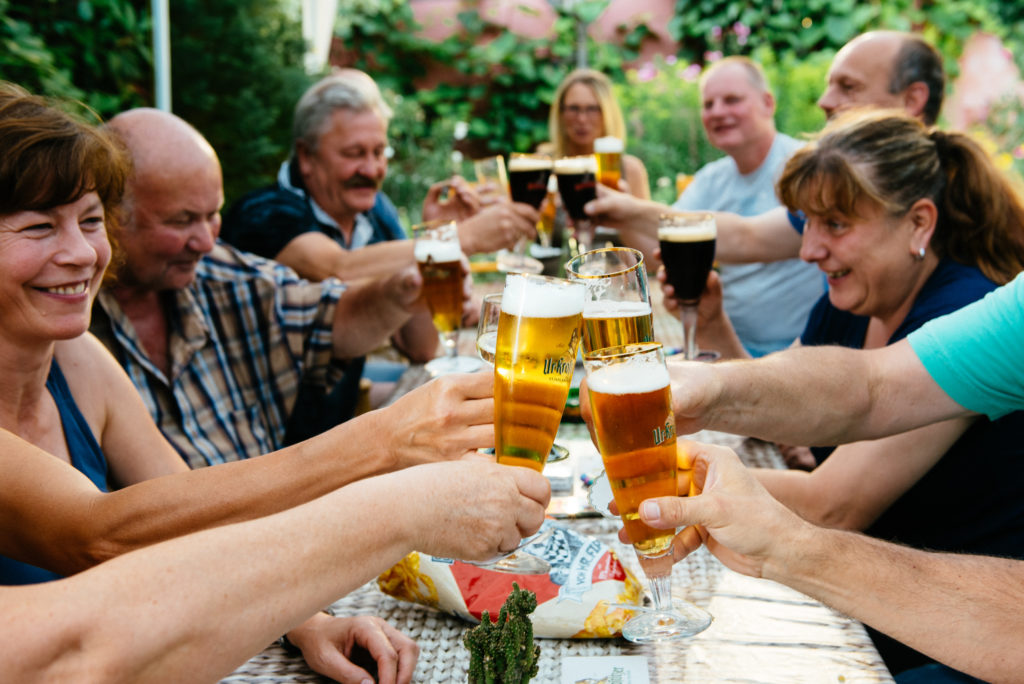 Mehrere Erwachsene stoßen an einem Gartentisch mit Biergläsern an. Sie lächeln und genießen die Gesellschaft.