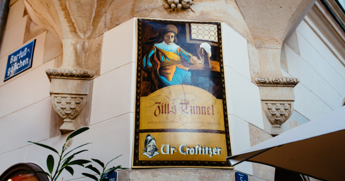 Ein gemaltes Porträt hängt an einer Steinsäule neben einem Straßenschild. Es zeigt eine Person in historischer Kleidung mit einem Getränk.