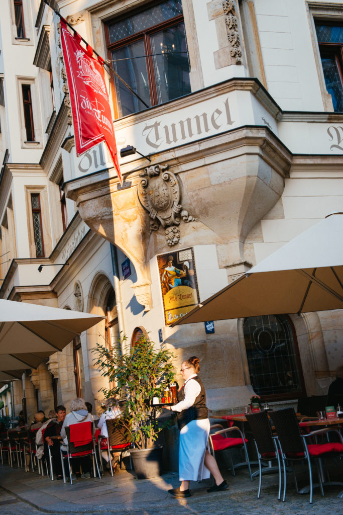 Ein Restaurant mit Außenbereich und Gästen; eine Kellnerin bedient. An der Fassade hängt eine rote Fahne.
