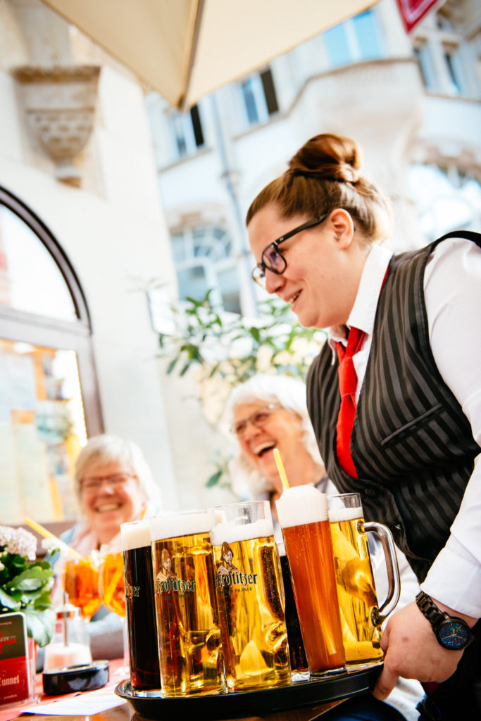Eine Kellnerin serviert lächelnd Bierkrüge an einem sonnigen Tag im Freien.