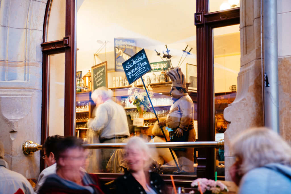 Personen sitzen in einem Café, ein Blick durch die offene Tür zeigt eine Figur mit Kochmütze.