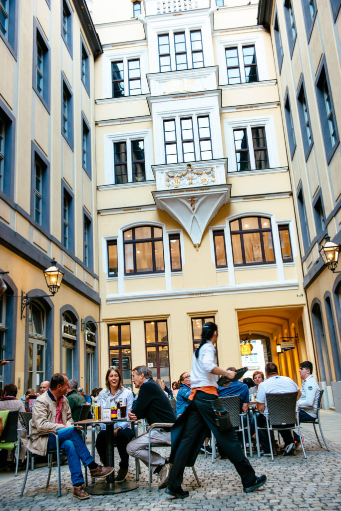 Menschen sitzen draußen an Tischen in einer belebten Gasse, Kellner geht vorbei.