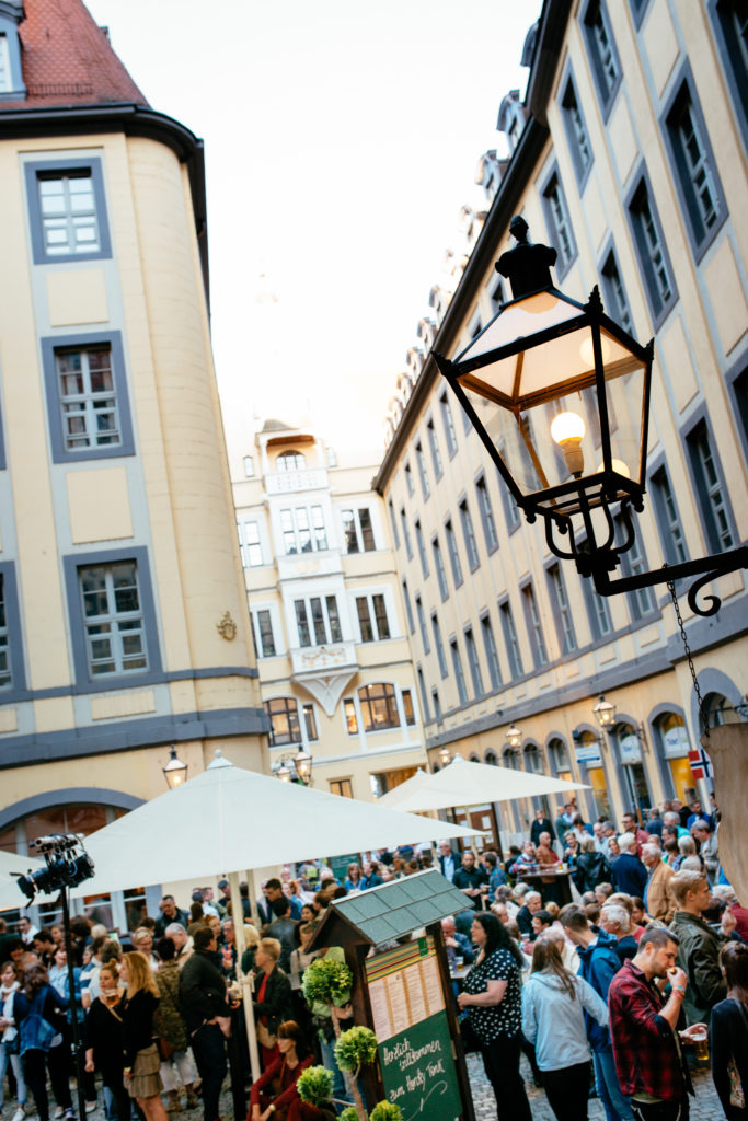 Ein belebter Marktplatz in einer Altstadt mit Menschen unter weißen Schirmen am Abend.