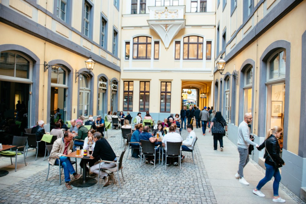 Menschen sitzen an Tischen in einer belebten Gasse, umgeben von historischen Gebäuden.