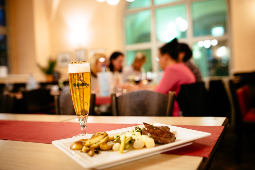 Ein Glas Bier steht im Vordergrund, dahinter ein Teller mit Essen auf einem Tisch in einem belebten Restaurant.