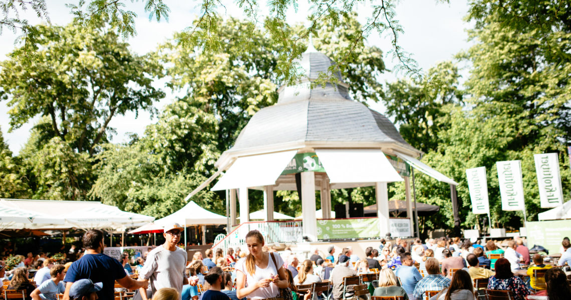 Viele Menschen sitzen draußen in einem Park bei einem Pavillon.