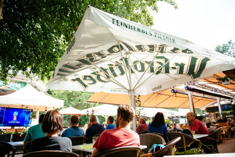 Menschen sitzen unter Sonnenschirmen und schauen auf einen Fernseher im Freien.