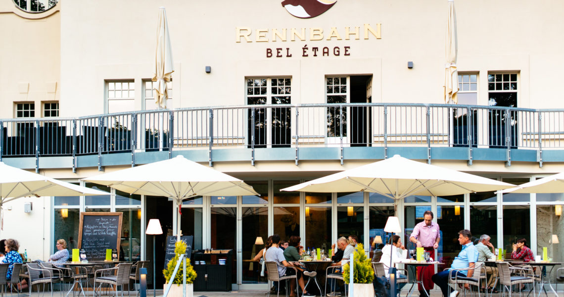 Mehrere Personen sitzen unter Sonnenschirmen vor einem großen Gebäude und speisen.