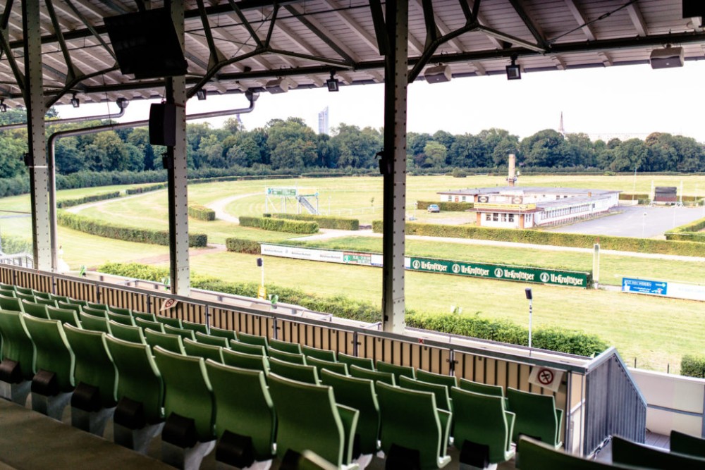Blick von einer überdachten Tribüne auf eine leere Pferderennbahn und grüne Sitzreihen.
