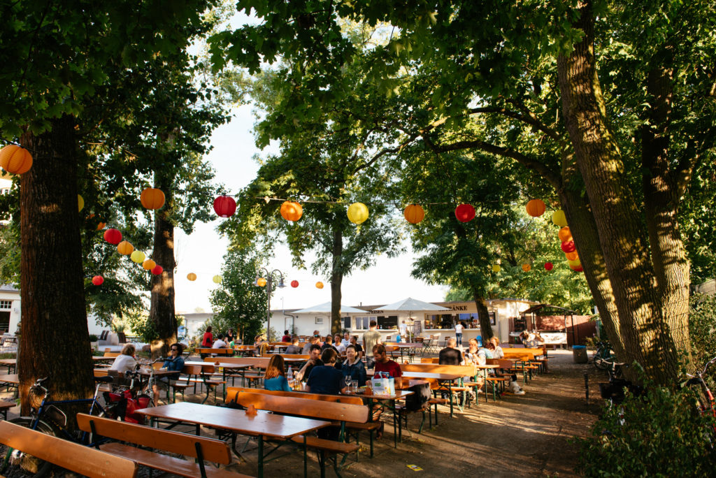 Ein belebter Biergarten im Freien mit bunten Lampions zwischen Bäumen und Gästen an Holztischen.