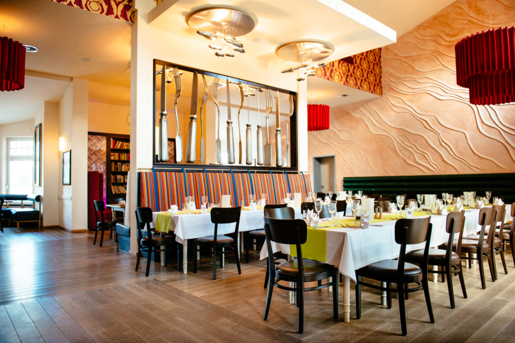 Ein helles Restaurant mit mehreren Tischen, farbigen Stühlen und kunstvoller Wandgestaltung.