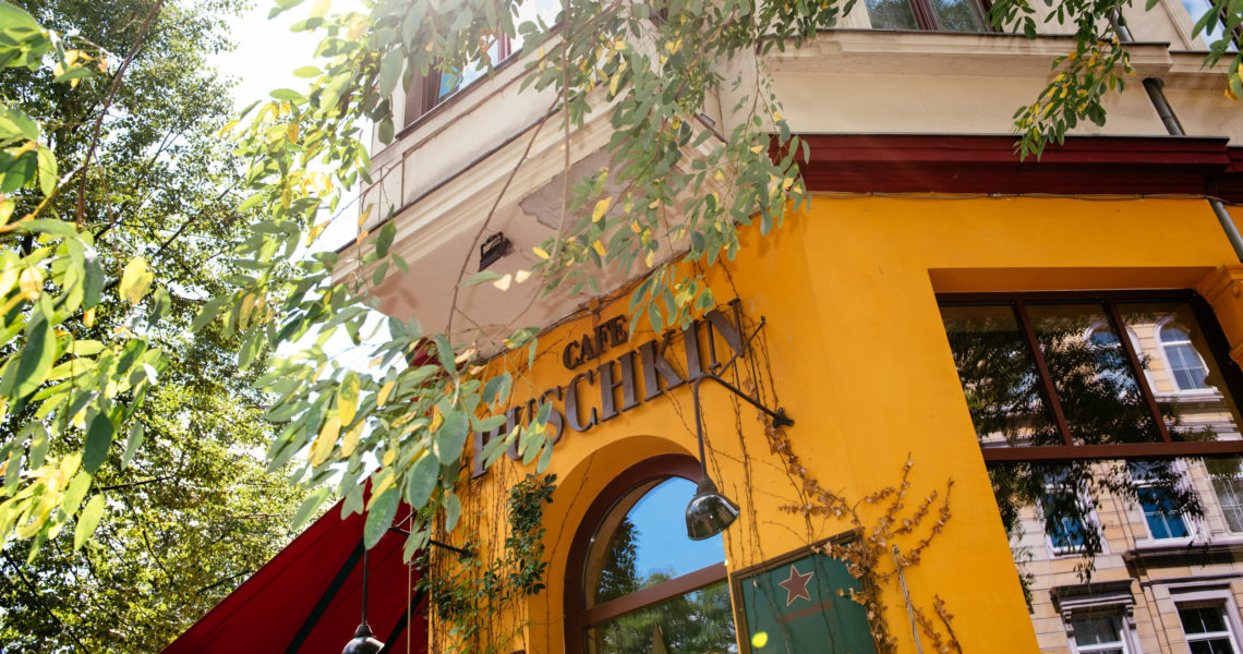 Sonniges gelbes Cafégebäude mit Bogeneingang und umgebenden grünen Blättern.