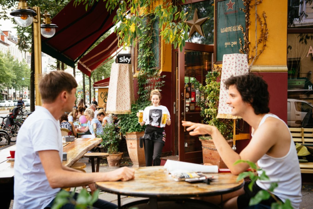 Ein Mann und eine Frau sitzen an einem Tisch in einem belebten Straßencafé und unterhalten sich. Eine Kellnerin nähert sich mit Getränken.