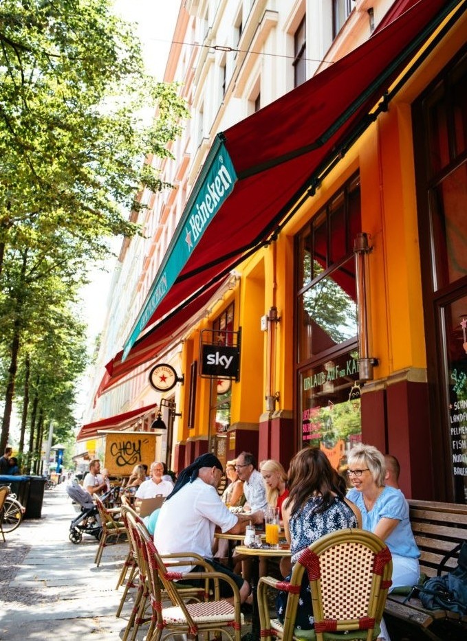 Menschen sitzen an Tischen auf einem Bürgersteig vor einem Café, umgeben von Bäumen.