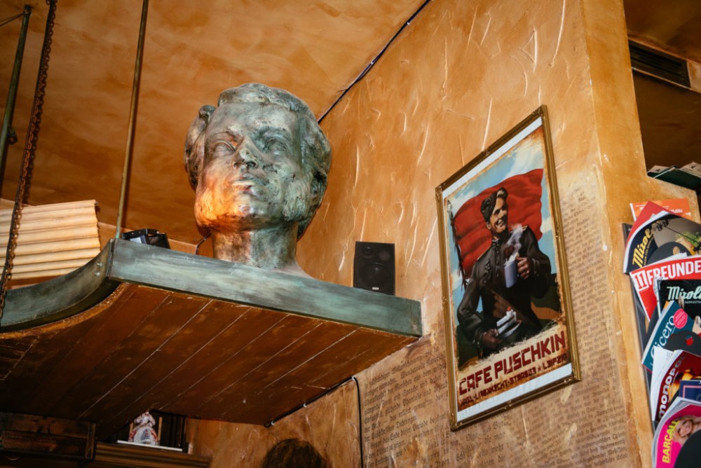 Eine große Büste steht auf einem Holzregal neben einem alten Poster an einer Wand.