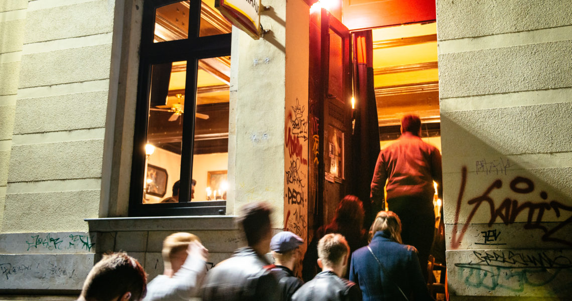 Menschen gehen bei Nacht an einem beleuchteten Lokaleingang vorbei. Graffiti ziert die Außenmauern des Gebäudes.