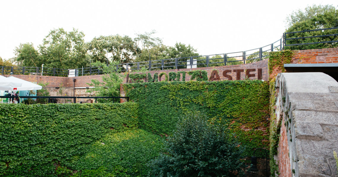 Eine bewachsene Ziegelmauer mit der Aufschrift aus Metallbuchstaben in einer Parklandschaft.