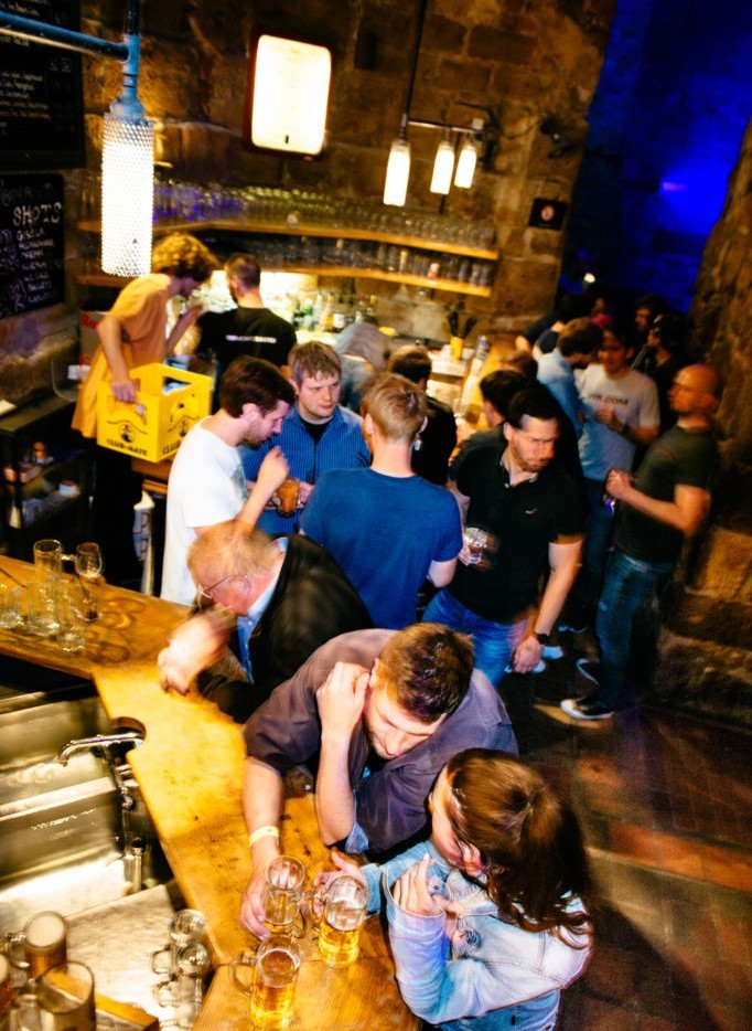 Menschen unterhalten sich an der Theke in einer belebten Bar. Einige stehen, andere sitzen mit Getränken.