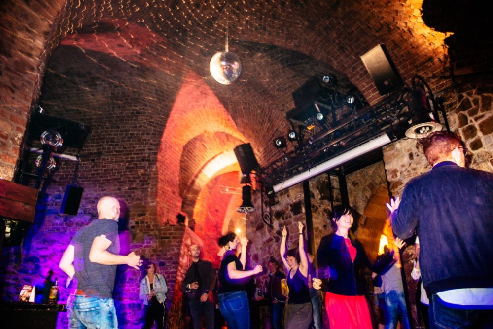 Personen tanzen in einem Raum mit gemauerten Gewölben und einer Diskokugel.