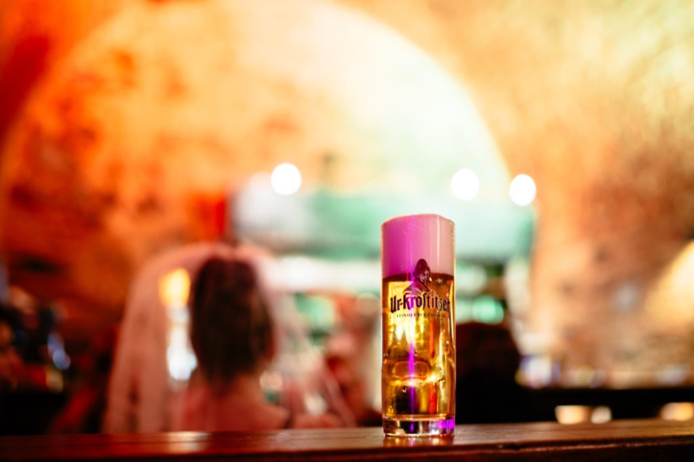 Ein Glas Bier steht im Vordergrund auf einer Bar, im Hintergrund sind unscharfe Personen.