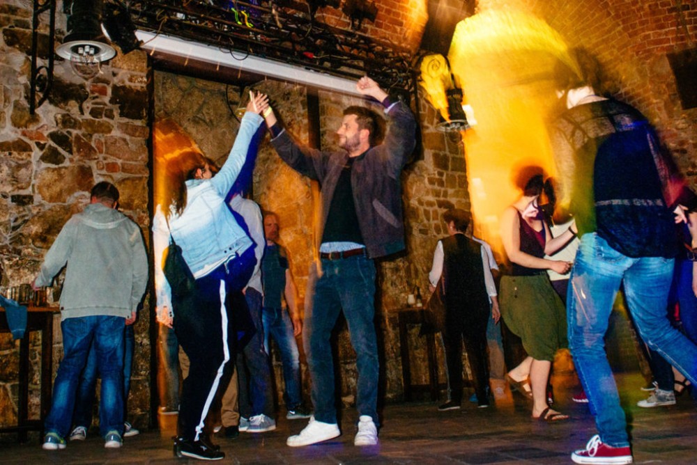 Mehrere Personen tanzen ausgelassen in einem Raum mit Steinwänden und bunten Lichtern.
