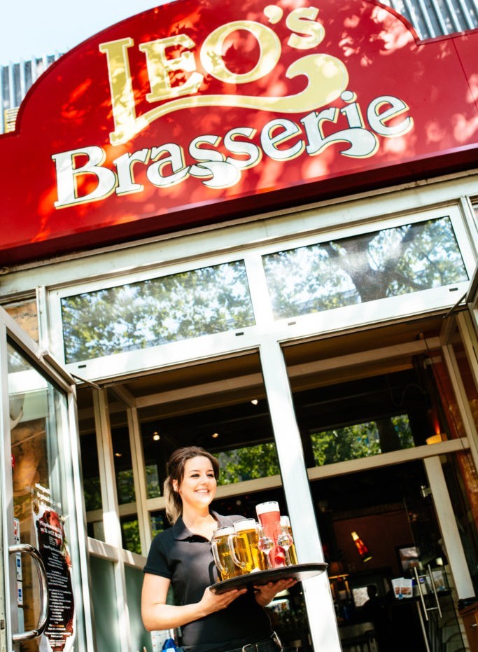 Eine lächelnde Kellnerin bringt Biergläser auf einem Tablett vor einem Restaurant.