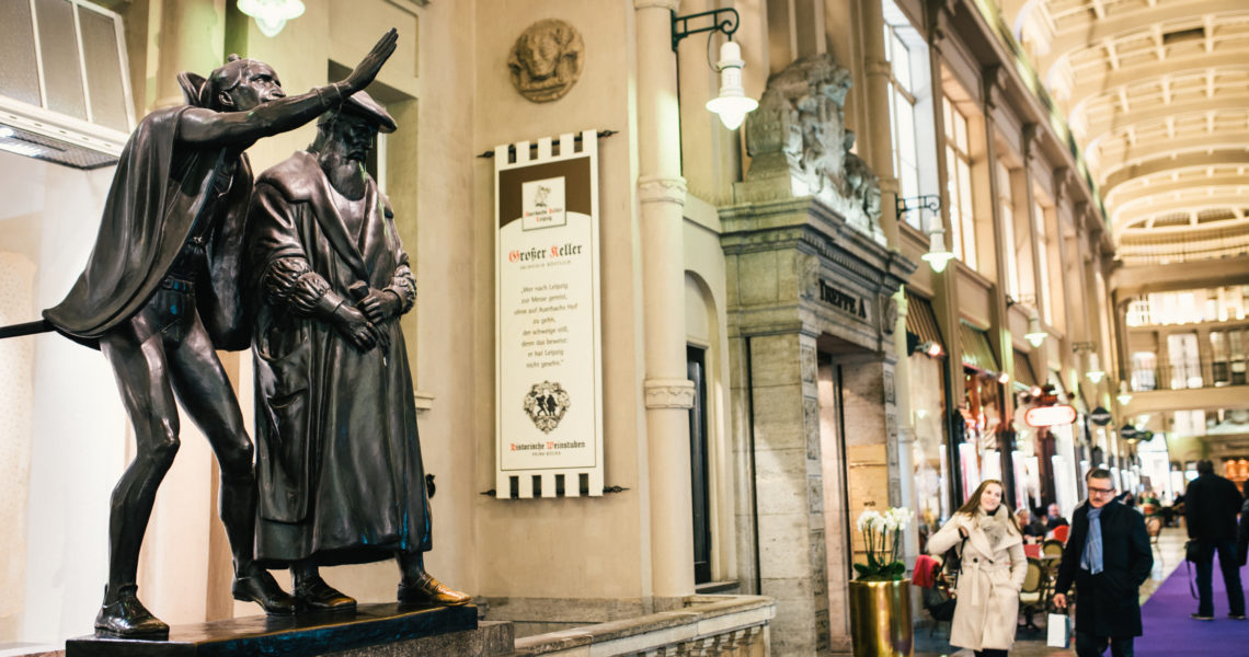 Zwei historische Skulpturen stehen vor einem belebten Einkaufspassage. Menschen gehen an ihnen vorbei.