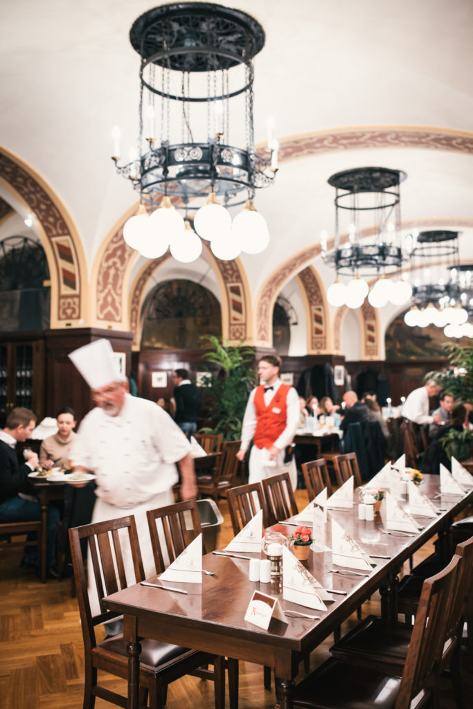 Ein Koch geht durch ein belebtes Restaurant mit elegantem Interieur und Gästen beim Speisen.
