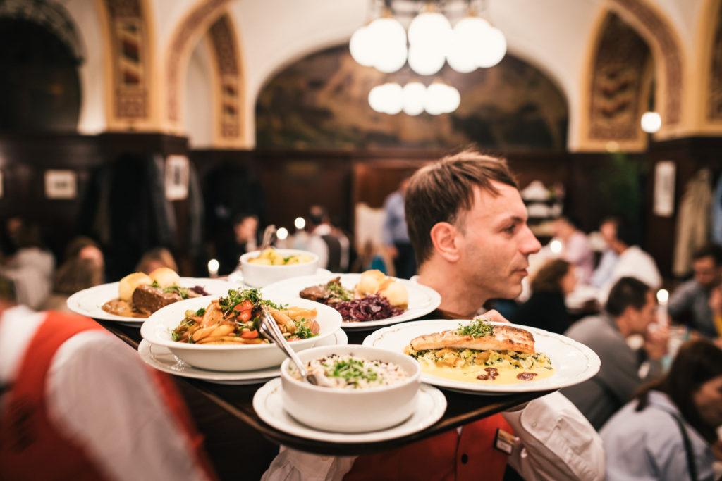 Ein Kellner trägt mehrere Teller mit verschiedenen Gerichten in einem belebten Restaurant.