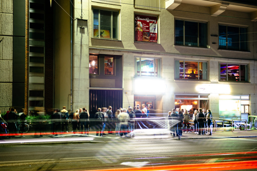 Eine Gruppe Menschen steht nachts vor einem beleuchteten Geschäft in einer Stadt.