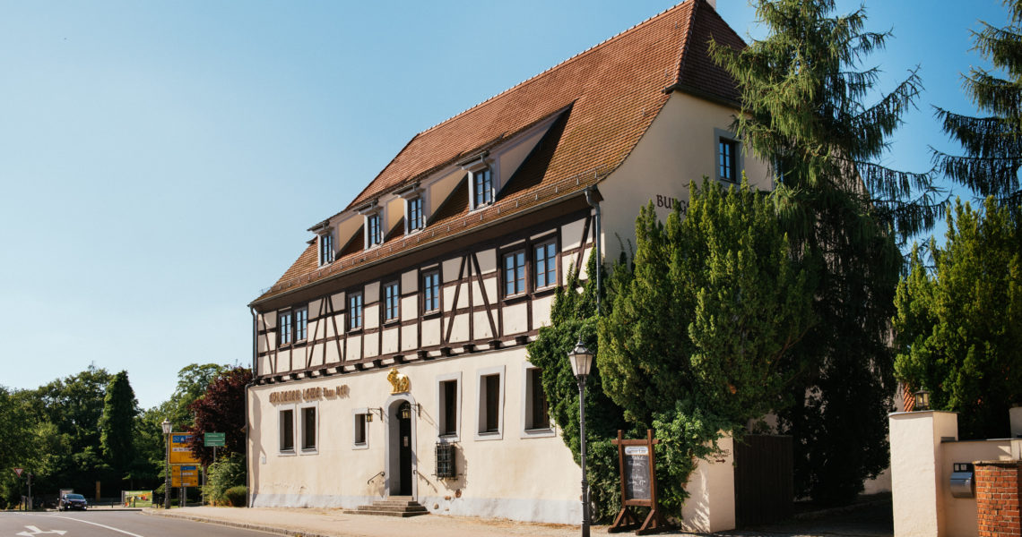 Ein Fachwerkhaus mit Satteldach steht sonnenbeschienen neben Bäumen.