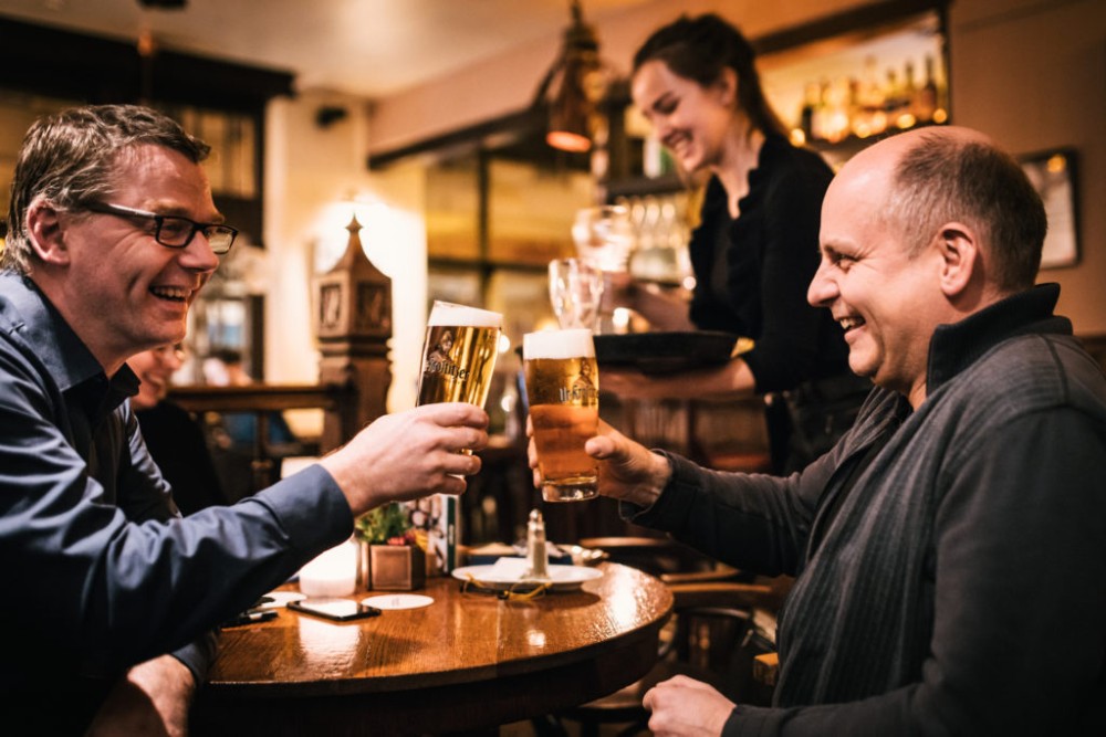 Zwei Männer stoßen in einem belebten Restaurant mit Bieren an. Sie lachen und genießen den Moment.
