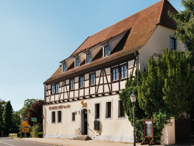 Traditionelles Fachwerkhaus mit rotem Ziegeldach und grünen Bäumen daneben.