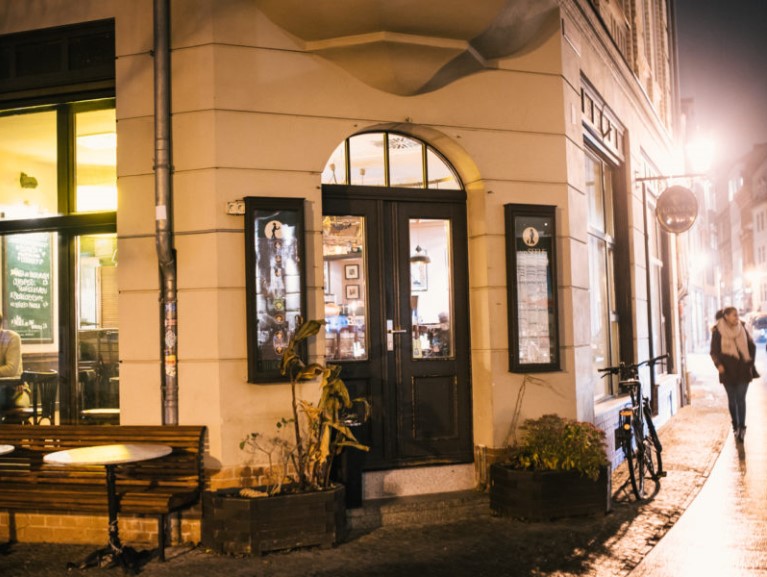 Eine beleuchtete Café-Ecke bei Nacht mit Passantin und parkendem Fahrrad.