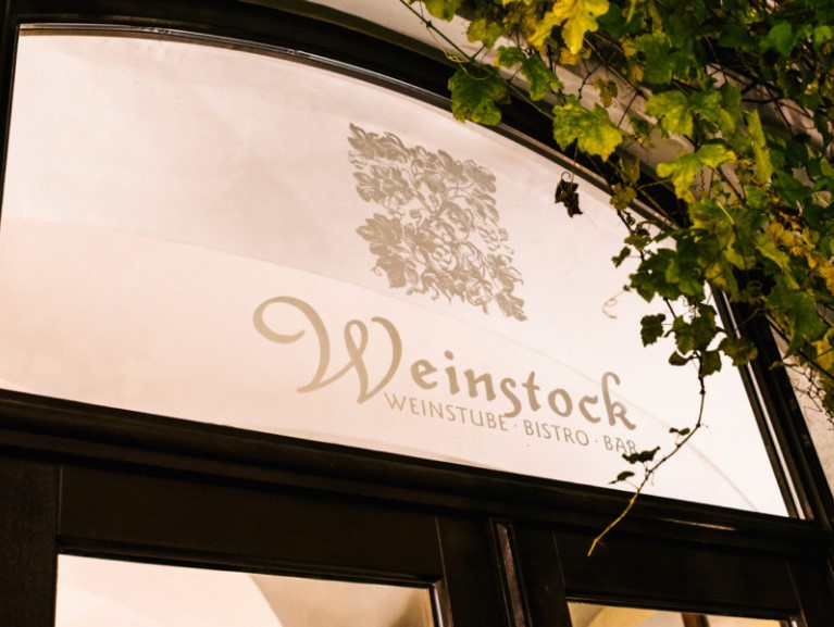 Ein Schild über einem Fenster zeigt einen stilisierten Weinstrauch und Schriftzüge.