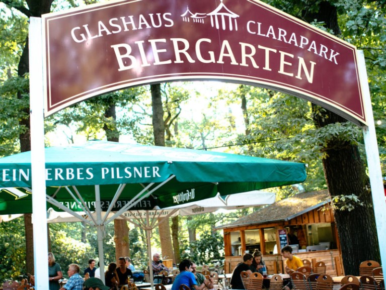 Ein Biergarten unter Bäumen mit Gästen an Holztischen, neben einem Getränkestand.