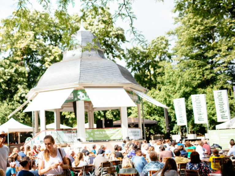 Viele Menschen sitzen in einem Park bei einem Pavillon und genießen das sonnige Wetter.