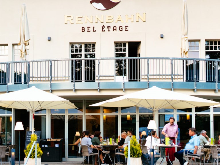 Menschen sitzen draußen in einem Restaurant unter Sonnenschirmen. Oben ist eine Terrasse sichtbar.