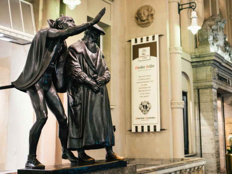 Zwei Bronzestatuen von Männern in historischen Gewändern stehen in einer belebten Halle.