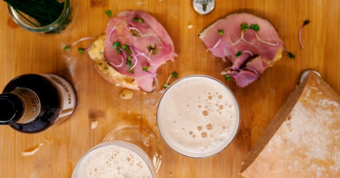 Zwei Gläser helles Bier stehen neben Tellern mit belegten Brötchen auf einem Holztisch.