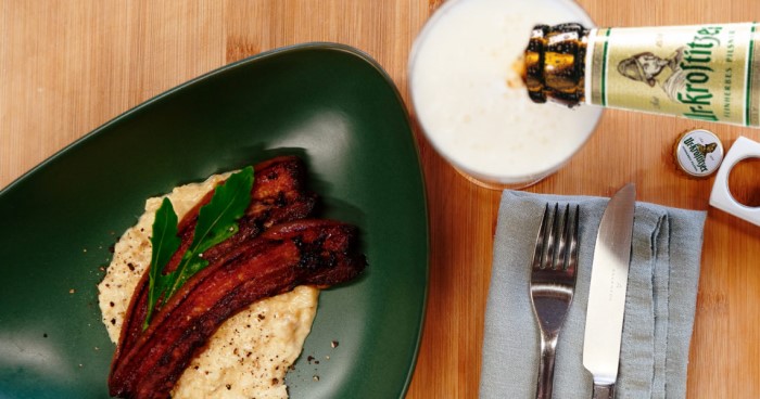 Gebratener Speck auf Hummus mit Rucola serviert auf einem grünen Teller, daneben Besteck und Bier.