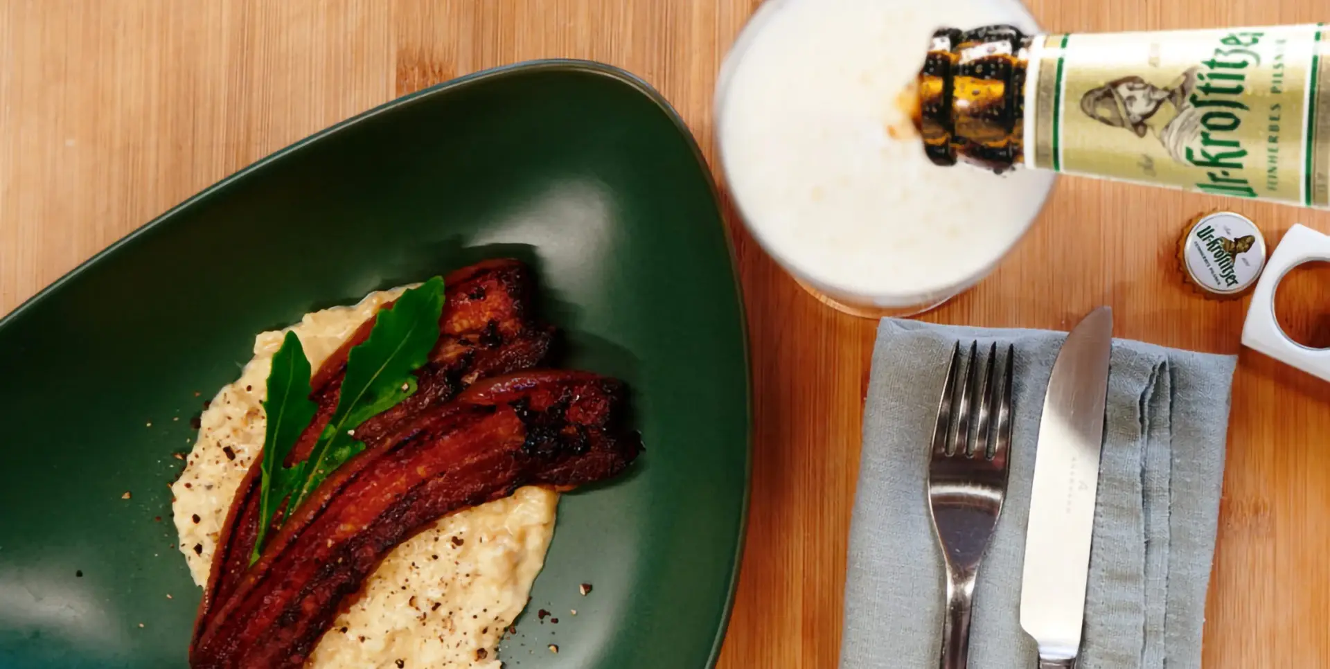 Auf einem grünen Teller liegt Speck auf einem Brei, neben einem Glas Bier und Besteck.